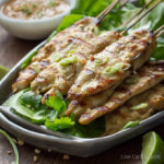 Grilled Thai chicken satay threaded on bamboo skewers on lettuce leaves on a gray plate served with peanut sauce and limes.