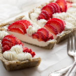 A closeup view of a sliced cream cheese and goat cheese strawberry tart in a press-in hemp and walnut crust with fresh strawberries arranged nicely among piped stars on top. The tart is on a piece of white parchment with silver forks in front to the side and a piece of white cheesecloth in the background.