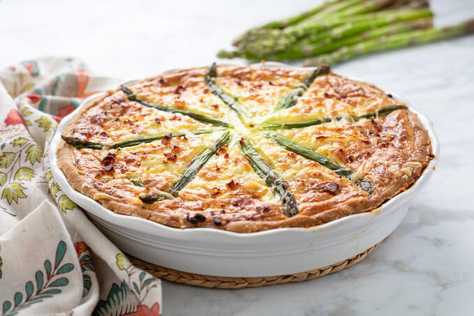 Ham and asparagus quiche in white ruffled pie plate with colorful napkin and asparagus spears.