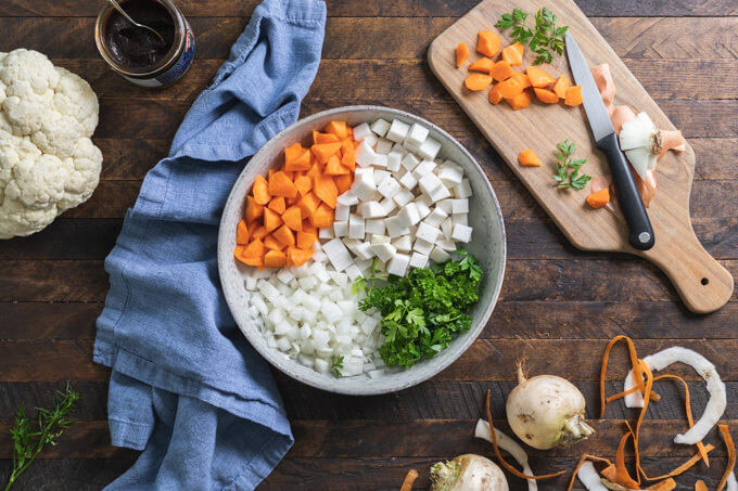 Keto cottage pie ingredients from left to right: cauliflower, beef base, bowl of diced carrot, turnip, onion and parsley. Cutting board with cut vegetables and knife.