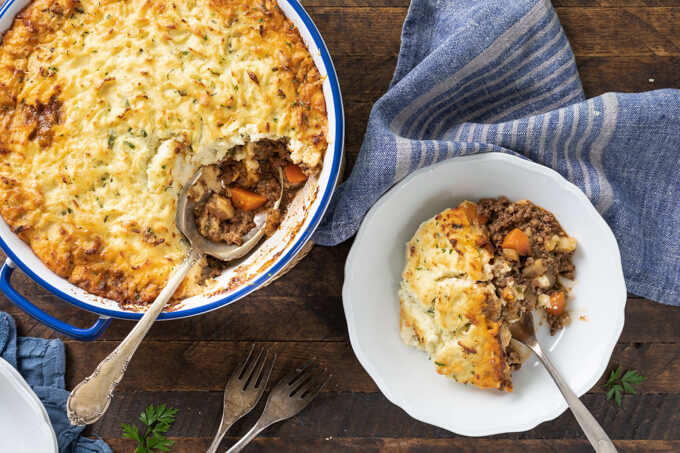 keto cottage pie in casserole dish with with serving spoon & individual serving in white bowl to the right with blue striped napkin.
